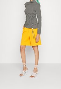 Černo-bílý kostkovaný top s dlouhým rukávem a nařasenou texturou, spárovaný se žlutými šortkami a bílými sandály na podpatku s pásky.