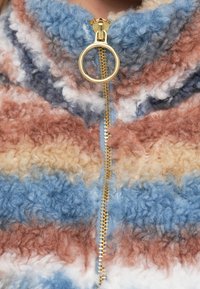 Chaqueta de forro polar con cierre de cremallera y rayas horizontales en marrón, azul y crema. Presenta una cremallera dorada, cuello alto y textura esponjosa.