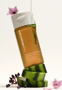 Brązowa szklana butelka toniku, biała nakrętka, zielony płyn, z purpurowym kwiatem na górze. Ułożona na plasterkach ogórka z ciemnymi jagodami w pobliżu.