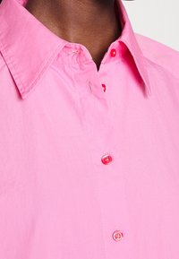 Rosa Baumwollhemd mit einem Kragen und roten Knöpfen, mit glatter Textur. Dargestellt vom Brustbereich aufwärts, mit Fokus auf die Knopfdetails.