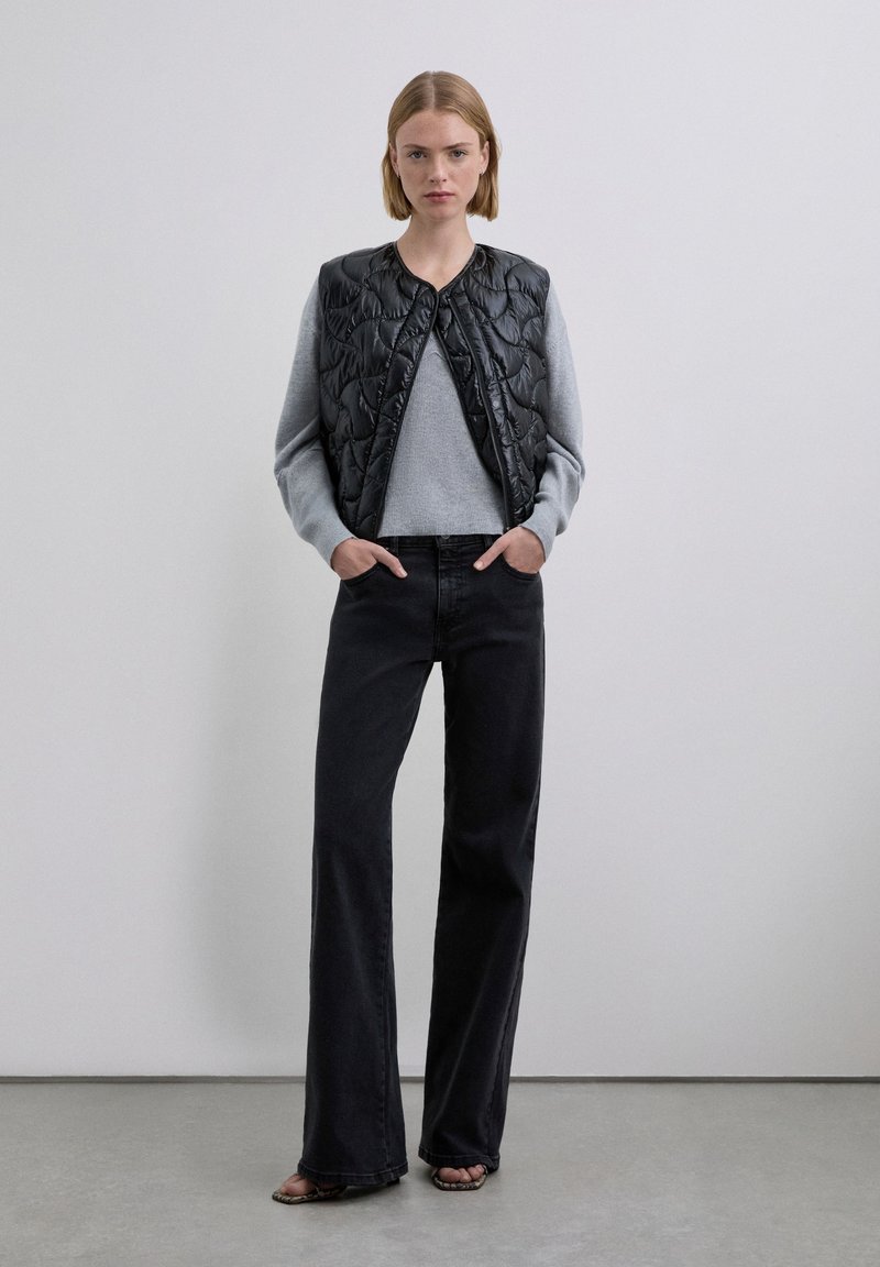 Black quilted vest over a light gray sweater, paired with high-waisted black jeans. Model stands with hands in pockets against a plain background.