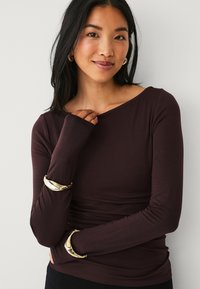 Haut à manches longues, marron foncé avec un décolleté bateau et une coupe ajustée ; il présente un tissu texturé et des bracelets dorés sur les deux bras.