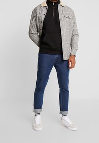 Rutig grå jacka med en krämfärgad krage över en svart zip-hoodie, i kombination med uppkavlade blå jeans och vita sneakers.