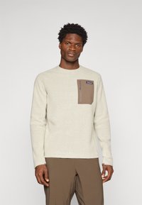 Pull-over en maille beige clair avec un motif texturé. Il est doté d'une poche poitrine marron avec une fermeture éclair et d'un logo de marque. Manches longues.