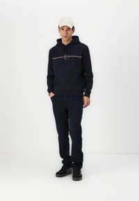 Navy hoodie with white and red horizontal stripe on chest, front pocket, adjustable drawstring hood, paired with dark jeans and black sneakers.