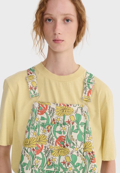 Jeune personne aux cheveux roux bouclés portant un t-shirt jaune pâle et une salopette à motifs floraux avec des dessins verts, jaunes et orange.
