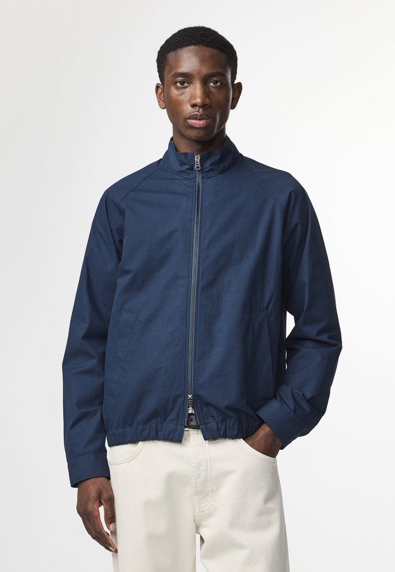 Mørkeblå zip-up jakke med høj krave, elastisk kant og forlommer. Glat stof med mat finish og raglan ærmer.