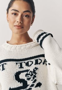 Suéter de punto grueso en color crema con estampado gráfico en azul marino, presenta un escote amplio y detalles a rayas en las mangas. Texturizado y acogedor.