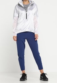 Chaqueta ligera plateada con media cremallera, mangas de malla; combinada con pantalones ajustados azul marino y zapatillas deportivas negras. Textura suave, corte moderno.