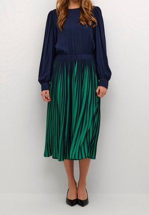Femme portant un chemisier bleu marine à manches longues et une jupe midi plissée verte avec des rayures noires, associée à des chaussures pointues noires.
