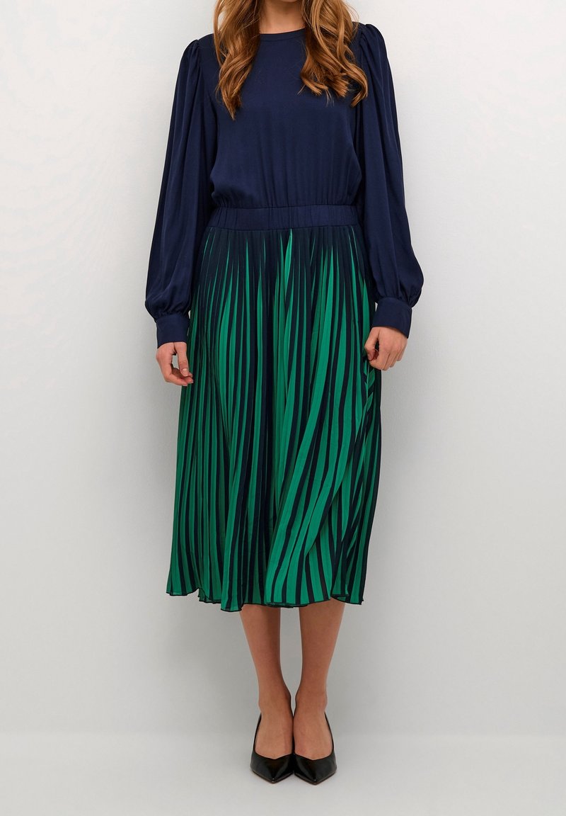 Femme portant un chemisier bleu marine à manches longues et une jupe midi plissée verte avec des rayures noires, associée à des chaussures pointues noires.