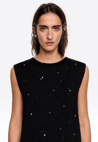 Czarny bezrękawnik wykonany z miękkiego materiału, ozdobiony rozmieszczonymi na przodzie i ramionach metalicznymi zdobieniami o różnych rozmiarach.