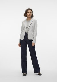 Světle šedý blazer s propracovaným designem, spárovaný se šedým topem a tmavě modrými širokými džínami. Hladká látka, přední kapsy a zapínání na jeden konec.
