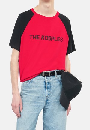 Person iført en rød og sort T-shirt med teksten "THE KOOPLES", lyseblå jeans med sort bælte, holder en sort baseballkasket ved siden.