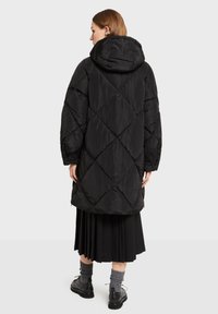 Sort puffert jakke med diamantformet quiltmønster, stor hætte og lange ærmer, båret over et plisseret sort skørt og grå sokker.