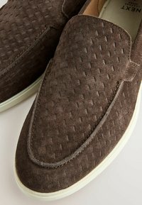 Chaussures en daim marron à enfiler avec une texture tissée et des semelles en caoutchouc blanc, étiquetées "NEXT" sur la doublure intérieure beige.
