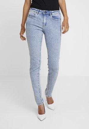 Žena nosící světle modré úzké džíny s vybledlým efektem a bílé špičaté vysoké podpatky, stojící před jednotným pozadím.