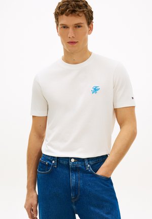 Giovane uomo che indossa una t-shirt bianca a maniche corte con monogramma blu e jeans blu, in piedi con una mano in tasca contro uno sfondo bianco.