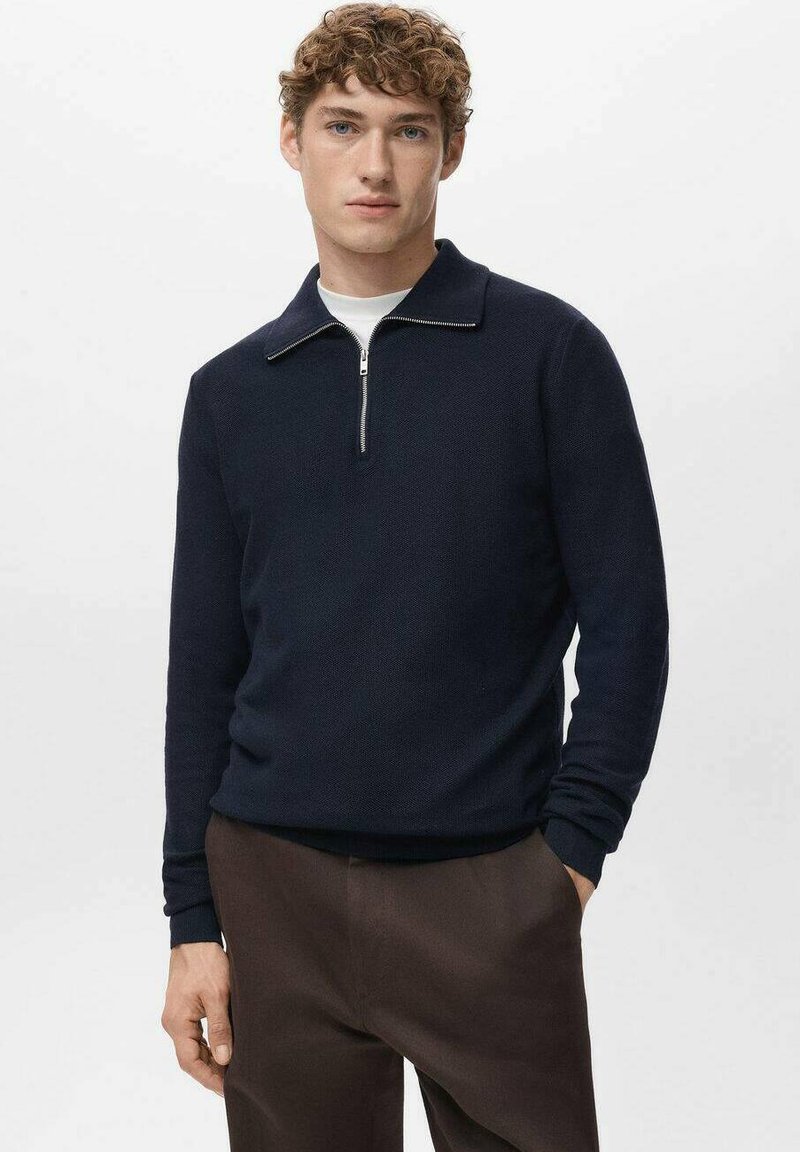 Pull-over en maille bleu marine avec col zippé, manches longues et tissu texturé. Porté sur une chemise blanche, associé à un pantalon marron.