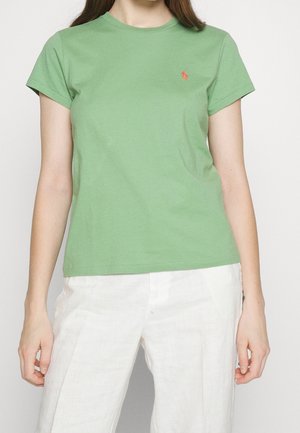 Femme portant un t-shirt vert uni à manches courtes avec un petit logo orange sur le côté gauche de la poitrine et un pantalon blanc, debout devant un fond uni.