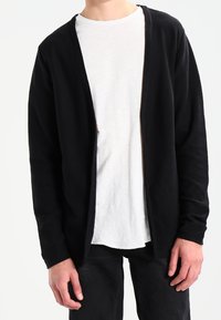 Person som bär en långärmad svart öppen cardigan över en vit t-shirt och svarta byxor, står med händerna avslappnat vid sidorna.