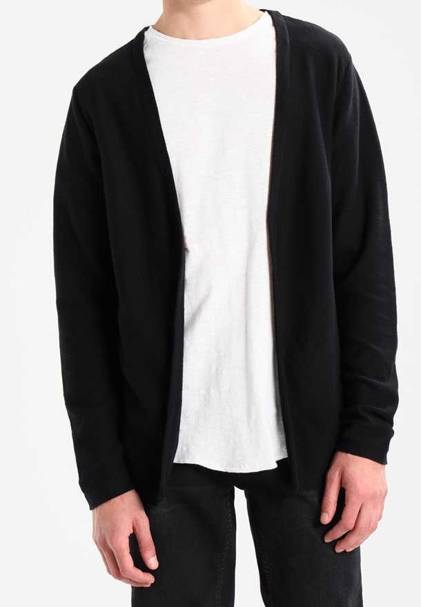 Person som bär en långärmad svart öppen cardigan över en vit t-shirt och svarta byxor, står med händerna avslappnat vid sidorna.