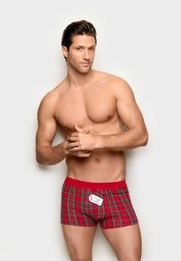 Czerwone szkockie bokserki w kratę z jednolitą czerwoną gumką w pasie; metka widoczna z przodu z informacjami o marce. Model z umięśnioną sylwetką, z założonymi rękami.