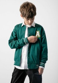 Zielona kurtka varsity z białymi akcentami, metalowymi zatrzaskami i haftowaną literą "A" na lewej piersi. Nałożona na białą koszulę z kołnierzykiem.