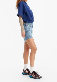 Blå oversize sweatshirt, ljusblå denimshorts med fransad nederkant, bruna och blå sportskor, och vita strumpor. Rutig långärmad tröja under.