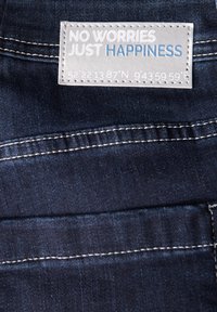 Tessuto denim di colore blu scuro con cuciture visibili. L'etichetta mostra la scritta "NESSUNA PREOCCUPAZIONE, SOLO FELICITÀ" e coordinate in grigio su uno sfondo chiaro.