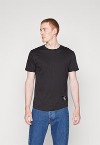 Black cotton t-shirt featuring a round neckline, short sleeves, and a logo patch on the left side. Paired with blue jeans.