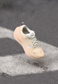 Adidași bej Nike pentru alergare, cu un superior din tricot texturat, logo-ul swoosh alb și o talpă de amortizare de culoare deschisă. Au șireturi și un tab de călcâi.