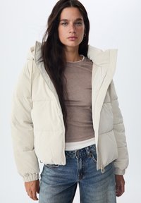 Doudoune beige clair avec capuche, dotée d'une fermeture éclair à l'avant, de poignets élastiques et d'une texture lisse, associée à un haut marron ajusté et un jean bleu.