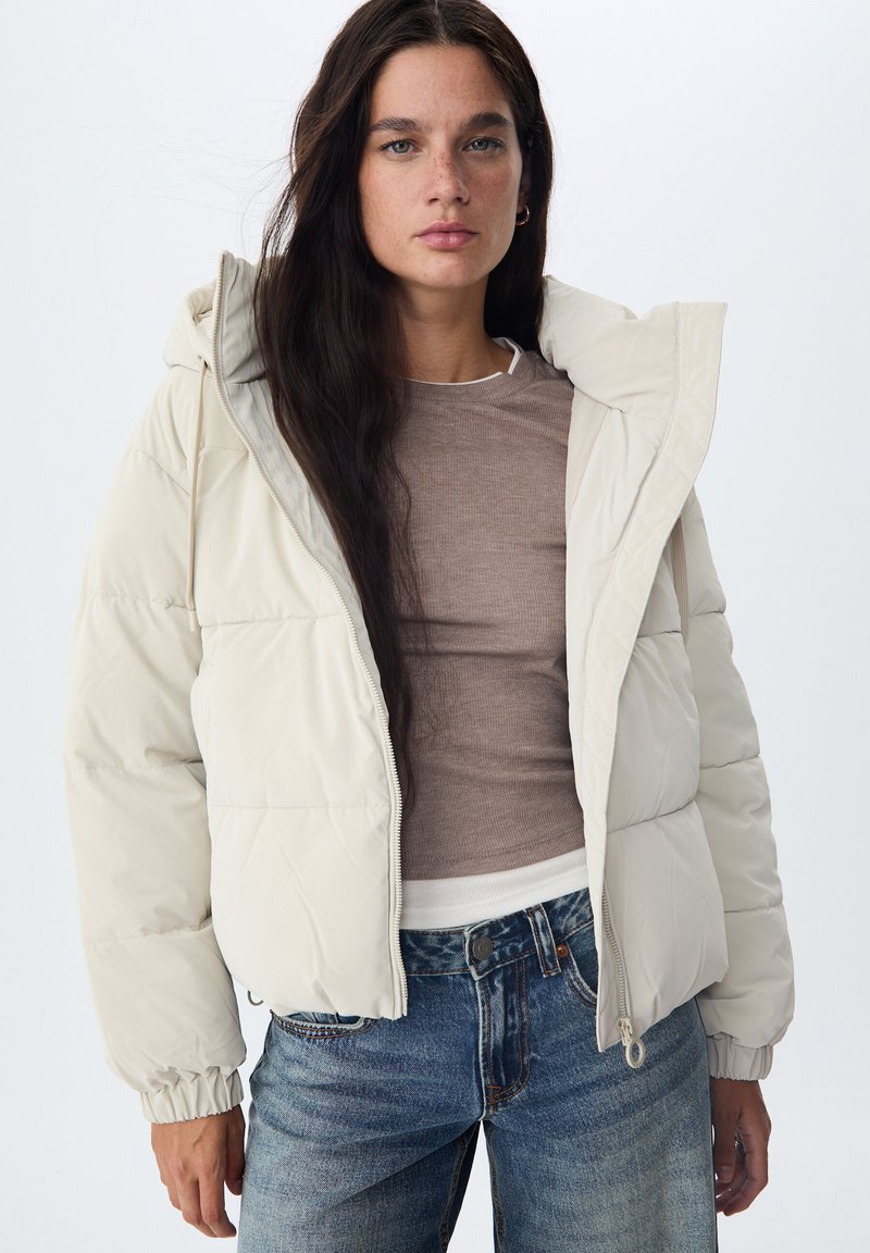 Doudoune beige clair avec capuche, dotée d'une fermeture éclair à l'avant, de poignets élastiques et d'une texture lisse, associée à un haut marron ajusté et un jean bleu.