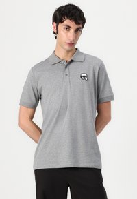 Siva polo majica iz mešanice bombaža. Ima ovratnik, kratke rokave in majhen črno-beli logotip na levem prsnem delu.