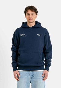Felpa blu navy, in misto cotone, con tasca frontale, cappuccio con lacci, logo bianco sul petto. Il modello indossa jeans blu chiaro.