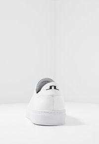 Bílé tenisky s hladkým kožním povrchem, zaobleným tvarem a černým logem na patě. Mají strukturovanou bílou gumovou podrážku a polstrovaný límec.