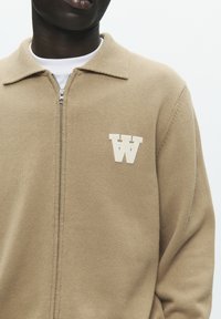 Cardigan beige à fermeture éclair avec col, texture côtelée, et un logo "W" beige sur la poitrine. Comprend une coupe standard et un tissu lisse.