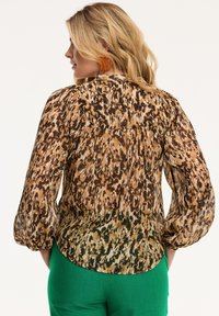 Blusa trasparente con un design a stampa animale marrone e beige, schiena plissettata e maniche a palloncino, abbinata a pantaloni verdi.