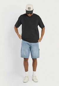 Camiseta de algodón negra con mangas cortas y un bolsillo en el pecho, combinada con pantalones cortos de mezclilla azul claro. Zapatillas blancas y una gorra beige con acentos negros.