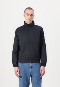 Jachetă ușoară neagră, cu fermoar în față, guler înalt și mansete elastice. Purtați peste pantaloni din denim albastru cu detalii de buzunar tip cargo.