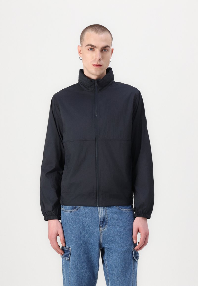 Must must jacket, mille ette, kõrge kraega ja elastsete varrukatega. Seljas sinised denim püksid, millel on tasku detail.