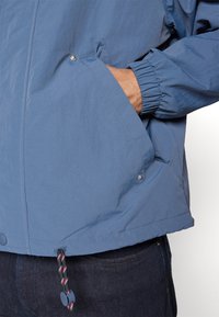 Light blue jacket featuring elastic cuffs, side pockets with snap buttons, and adjustable drawstring hem. Made of smooth, lightweight fabric.