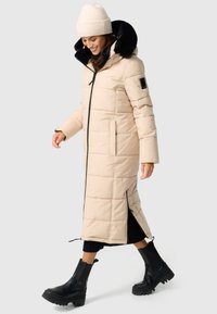 Beige langer Puffer-Mantel mit Kapuze aus schwarzem Kunstfell, Seitentaschen und Reißverschlüssen am Saum, kombiniert mit schwarzen Stiefeletten.