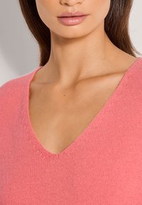 Rosa gestrickter Pullover mit V-Ausschnitt, der eine strukturierte Oberfläche und dezente gewellte Nähte entlang der Kanten aufweist.