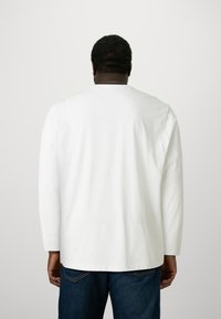 Man med kort hår som bär en enkel vit långärmad skjorta och blå jeans, stående vänd bort från kameran mot en neutral bakgrund.