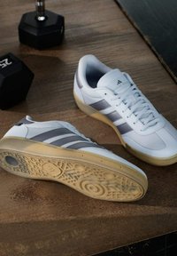 Witte sportieve schoenen met grijze strepen, textielstof en rubberen zolen. Geplaatst op een houten oppervlak naast een zwarte dumbbell.