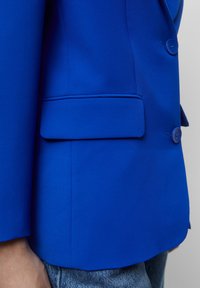 Blazer bleu présentant une coupe ajustée, une texture lisse, une poche poitrine unique et des boutons ronds, posé sur un fond gris clair.