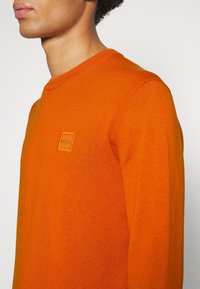 Oranssi neulepaita, jossa on pyöreä kaula-aukko, ribattuja hihansuita sekä pieni, teksturoitu BOSS-logo-etiketti rinnassa. Pehmeä neulemateriaali.