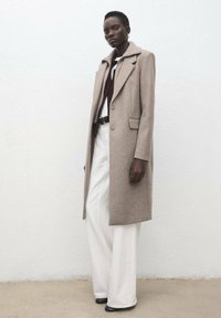 Cappotto di lana beige con colletto a rever e tre bottoni, indossato sopra una camicia color borgogna e pantaloni bianchi a gamba larga, abbinato a scarpe con tacco nere.